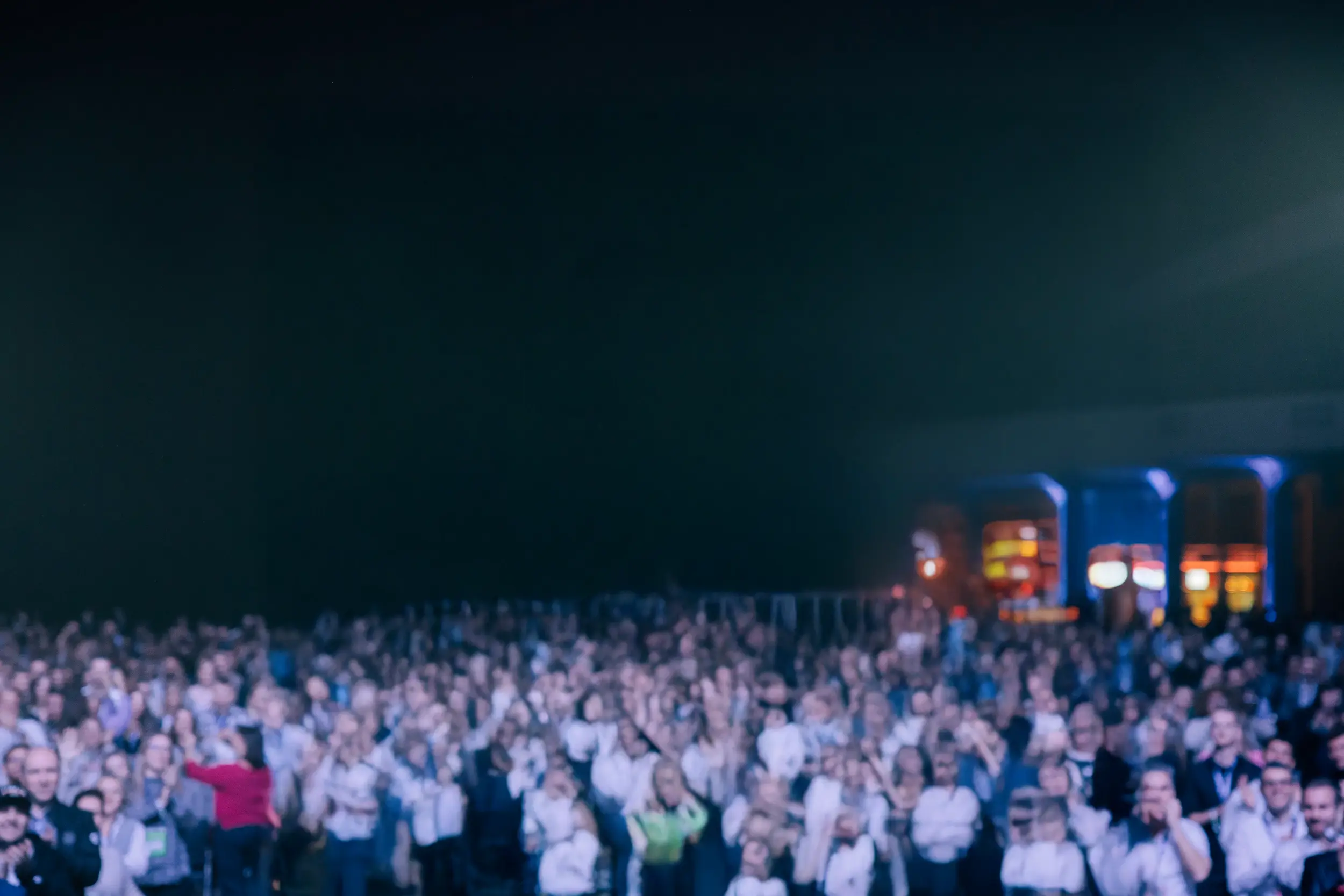 Large crowd of people gathered indoors, facing forward, with bright lights in the background.
