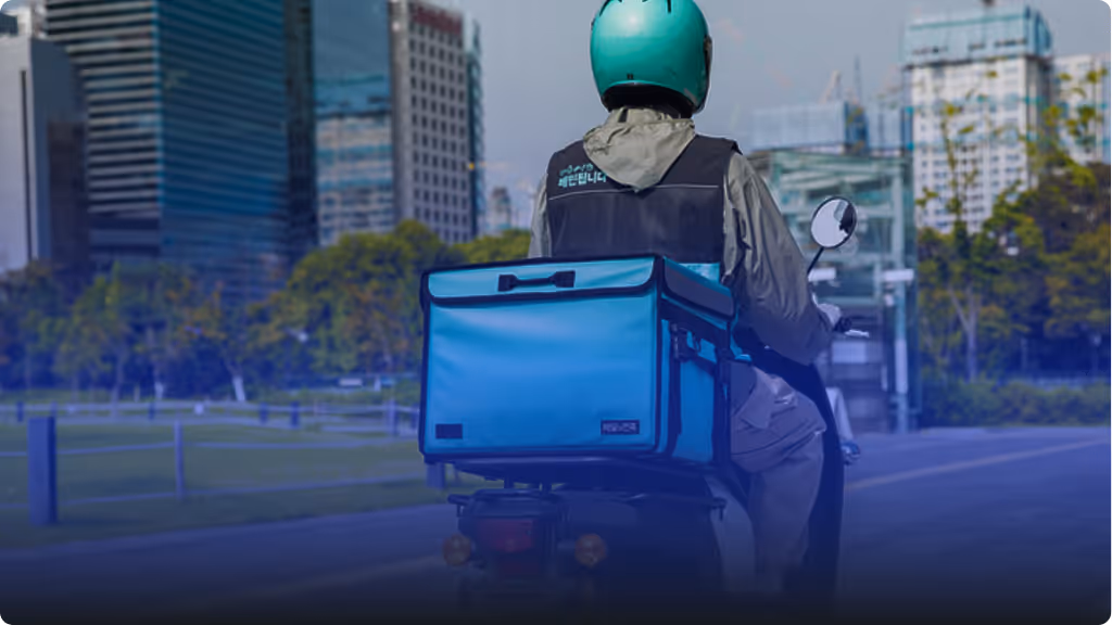 Delivery person wearing a green helmet riding a scooter with a large blue insulated delivery bag in an urban setting.