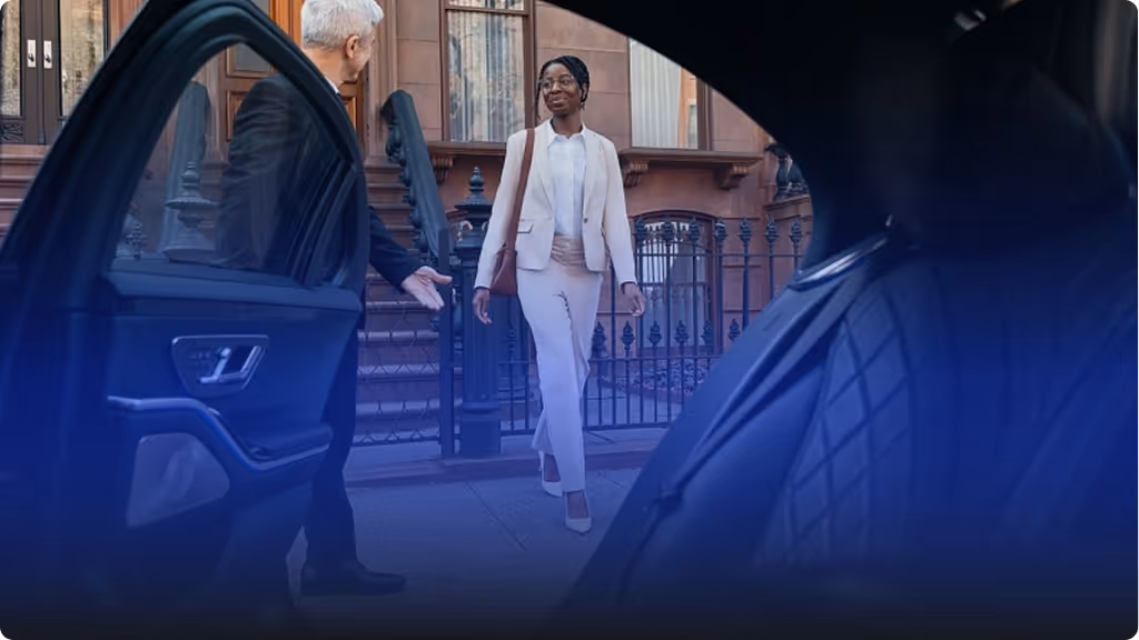 Man holding car door open for woman in business attire on city sidewalk near brownstone buildings.