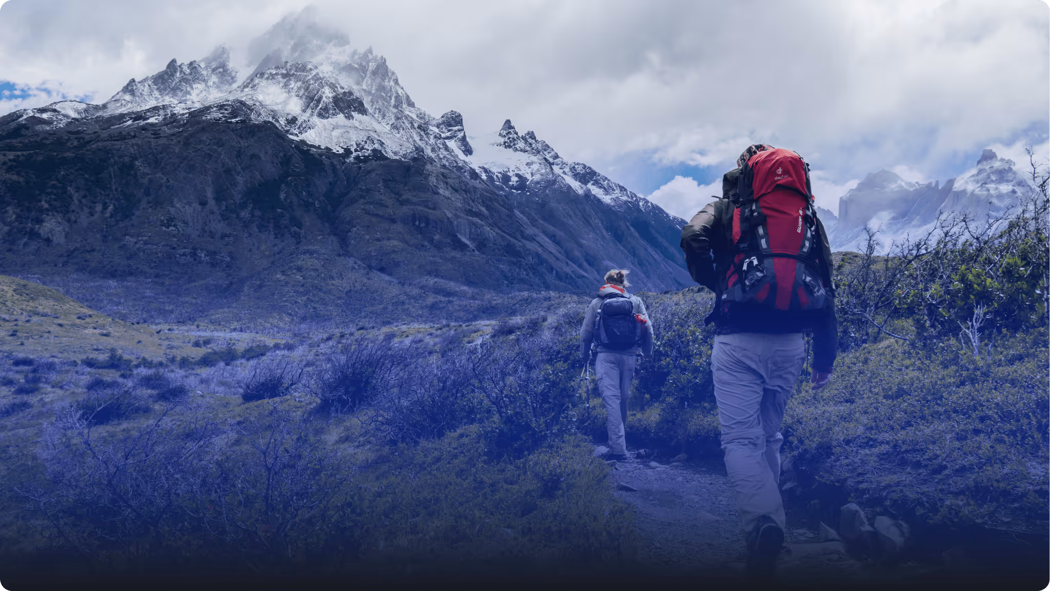 Two hikers with backpacks walking on a trail toward snowy mountain peaks under a cloudy sky.