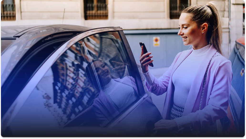 Woman holding phone and opening car door on a city street, smiling as she looks at the screen.