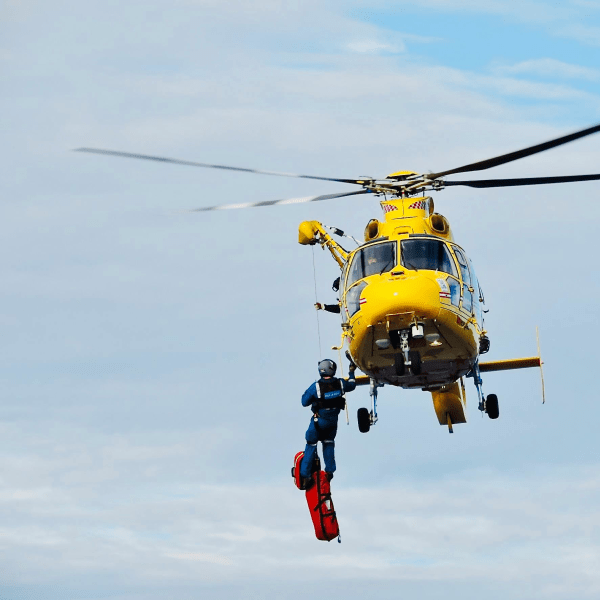 A yellow helicopter conducting a rescue mission with a crew member suspended below.