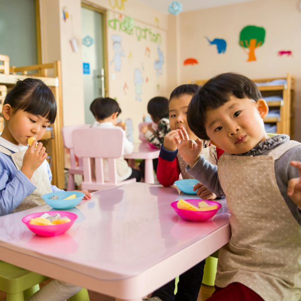 Kids seated around a table in a colorful classroom, eating snacks happily.