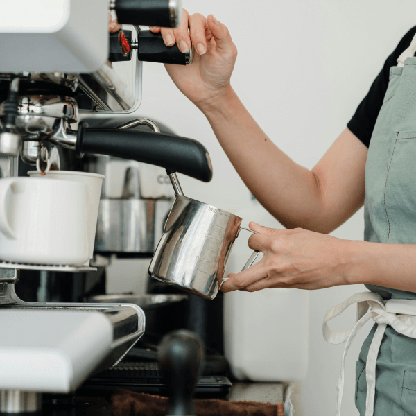 Crop faceless young female barista in apron steaming milk at modern coffee machine while preparing delicious latte