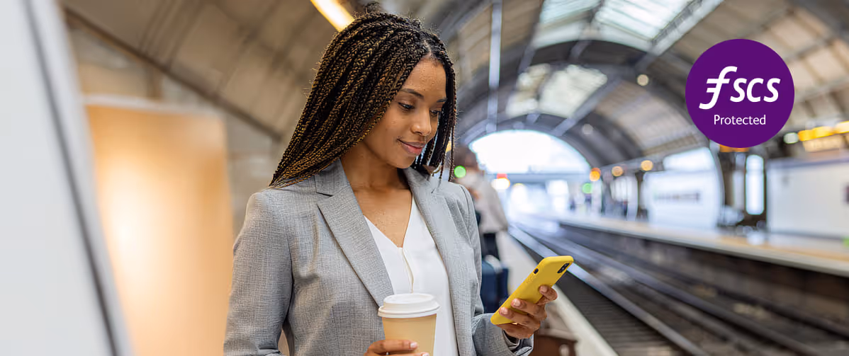 Woman in gray blazer holding a coffee cup and looking at her yellow smartphone on a train platform.