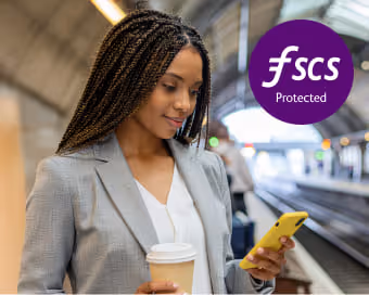 Woman in a blazer holding a coffee cup and looking at her yellow smartphone on a train platform with FSCS Protected logo.