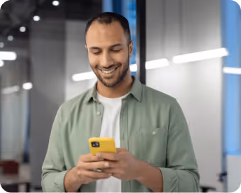 Smiling man in a green shirt looking at his yellow smartphone with an FSCS Protected logo in the top right corner.