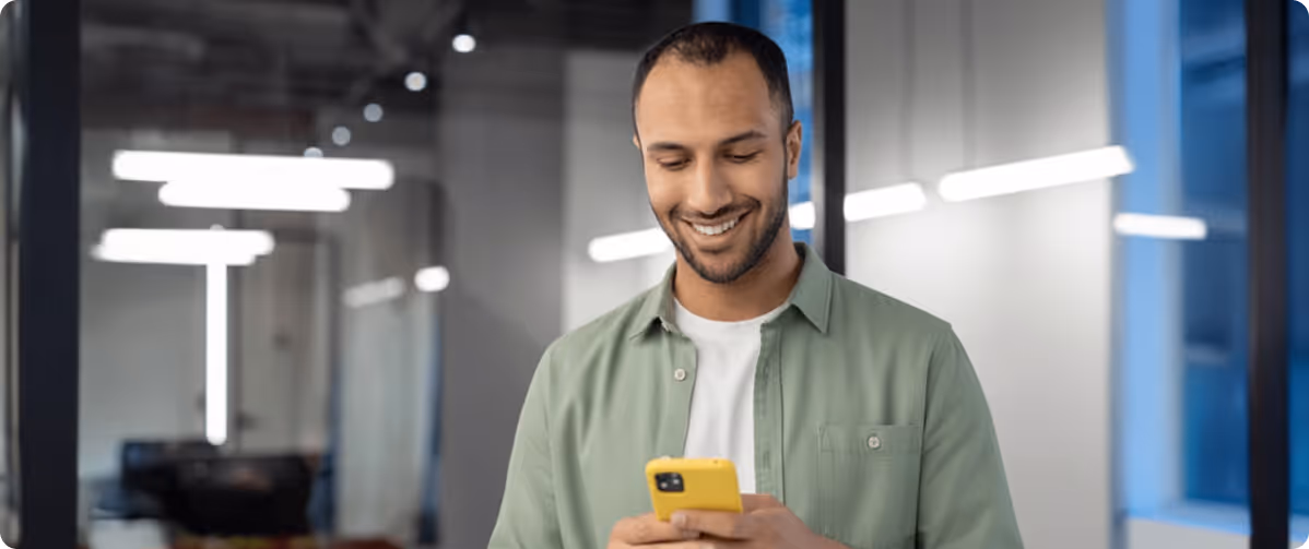 Smiling man in green shirt looking at his yellow smartphone inside a modern office with FSCS Protected logo.