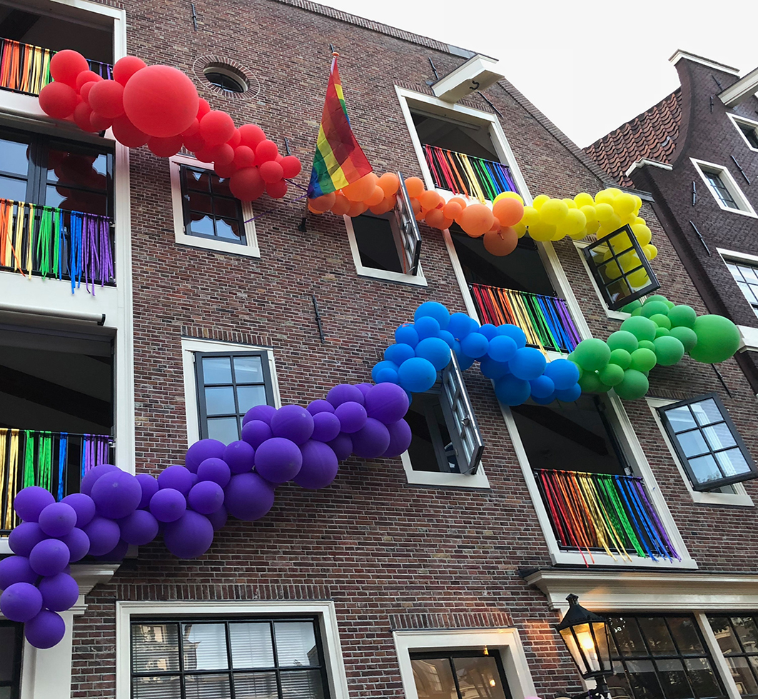 A brick building with a rainbow pride flag on the front and garlands of rainbow colored balloons spread across the face of the building.