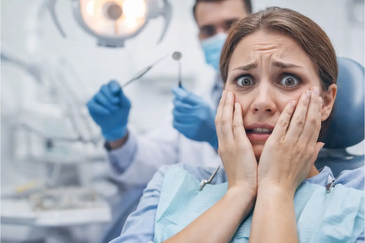 Anxious woman in a dentist's chair covering her mouth, looking scared as the dentist prepares tools.