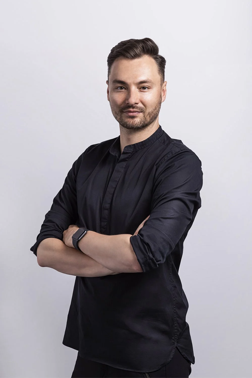 Man with short dark hair and beard wearing a black shirt, standing with arms crossed and smiling slightly against a light background.