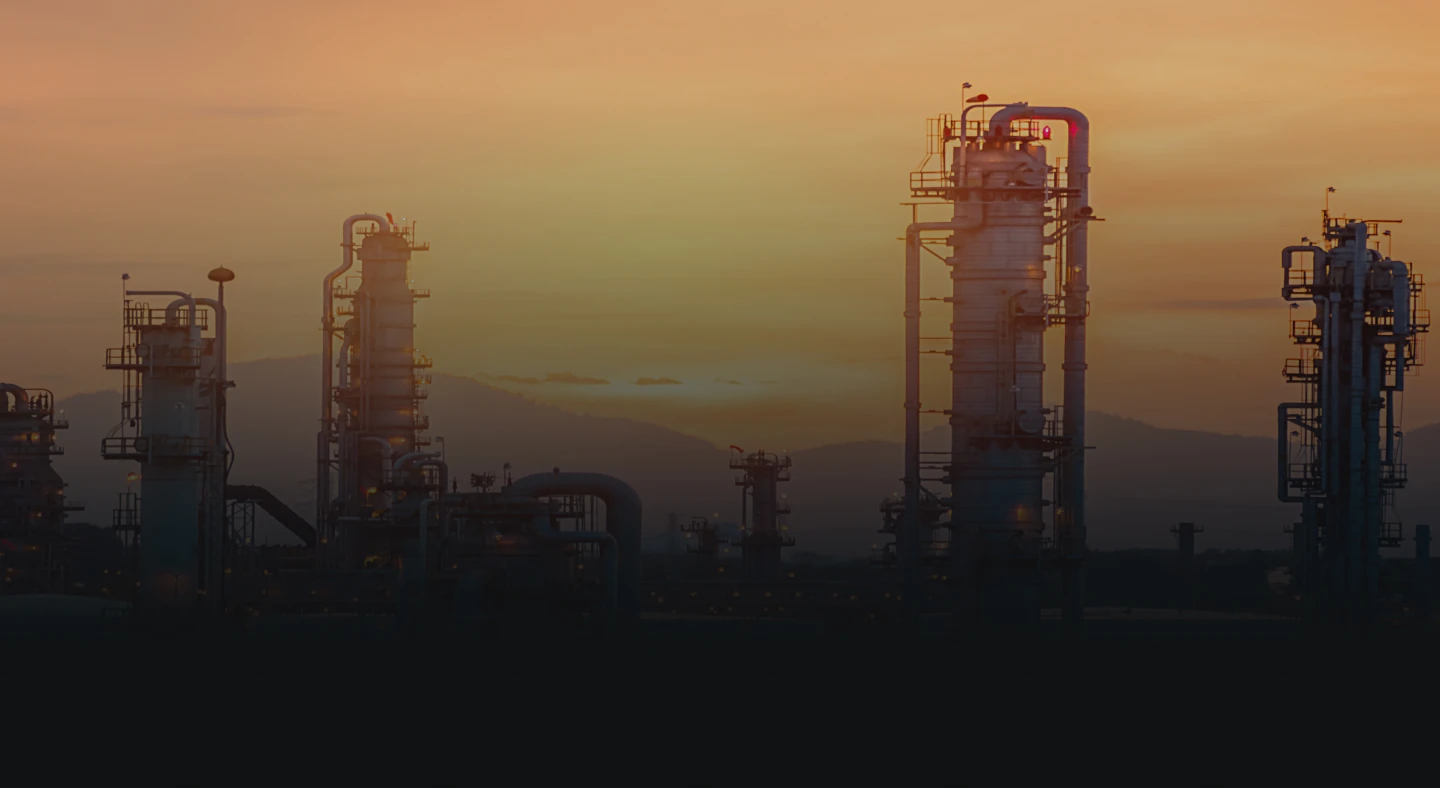 Silhouettes of industrial chemical plant towers set against an orange sunset sky with distant hills.
