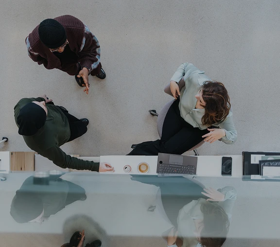 Overhead view of three people at a desk with a laptop, coffee cup, and smartphone.