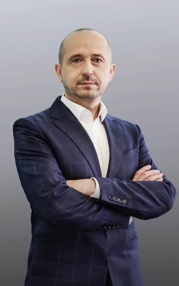 Confident man with short hair wearing a navy checkered suit and white shirt standing with arms crossed against a gray gradient background.