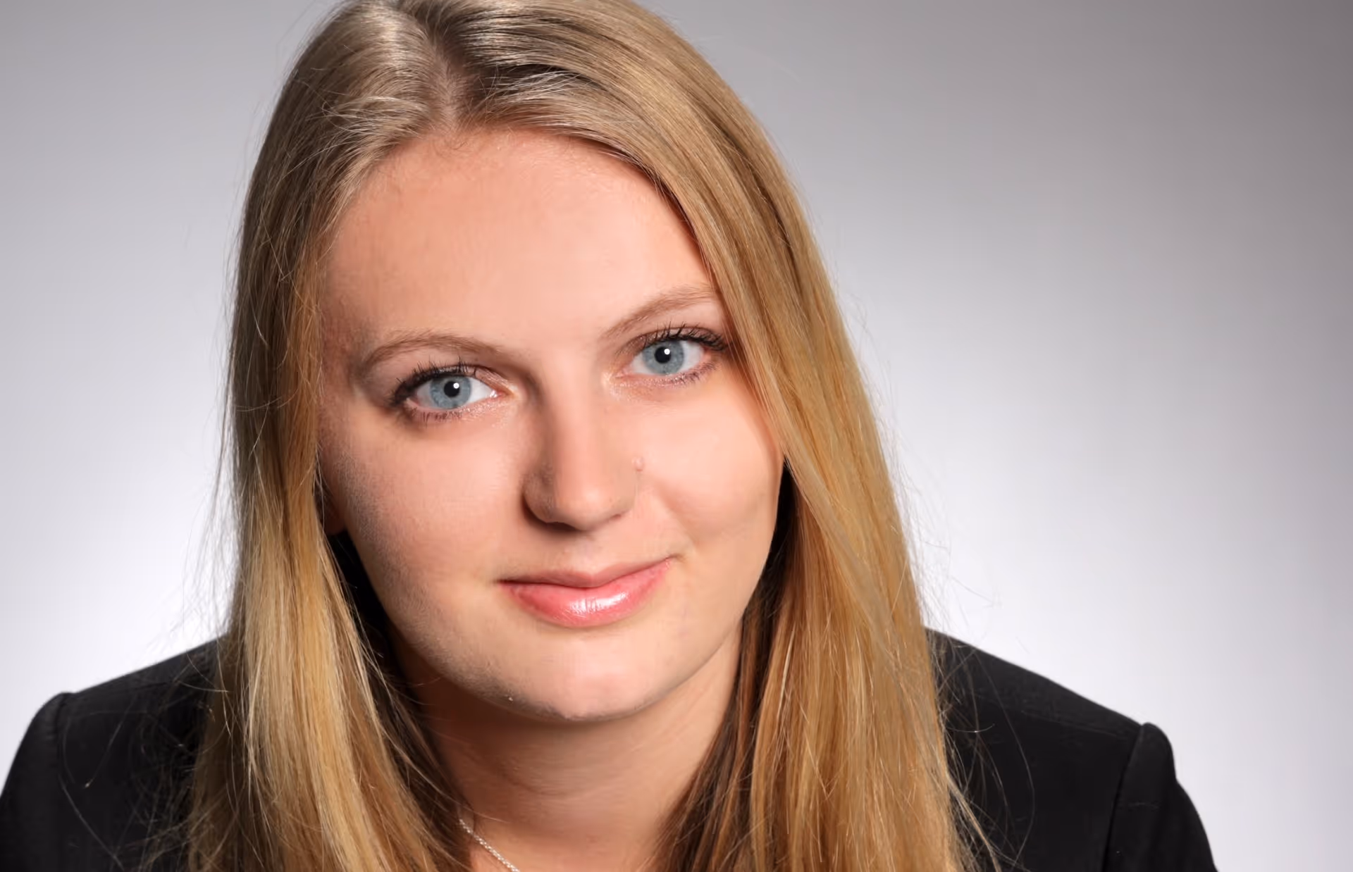 Close-up portrait of a woman with long blonde hair, blue eyes, and a slight smile, wearing a black jacket.