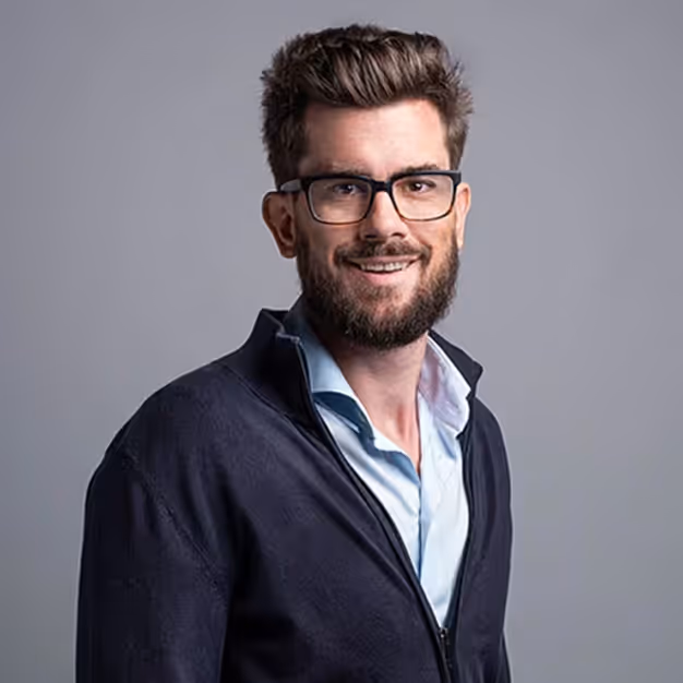 Smiling man with dark hair, beard, and glasses wearing a light blue shirt and dark jacket against a gray background.