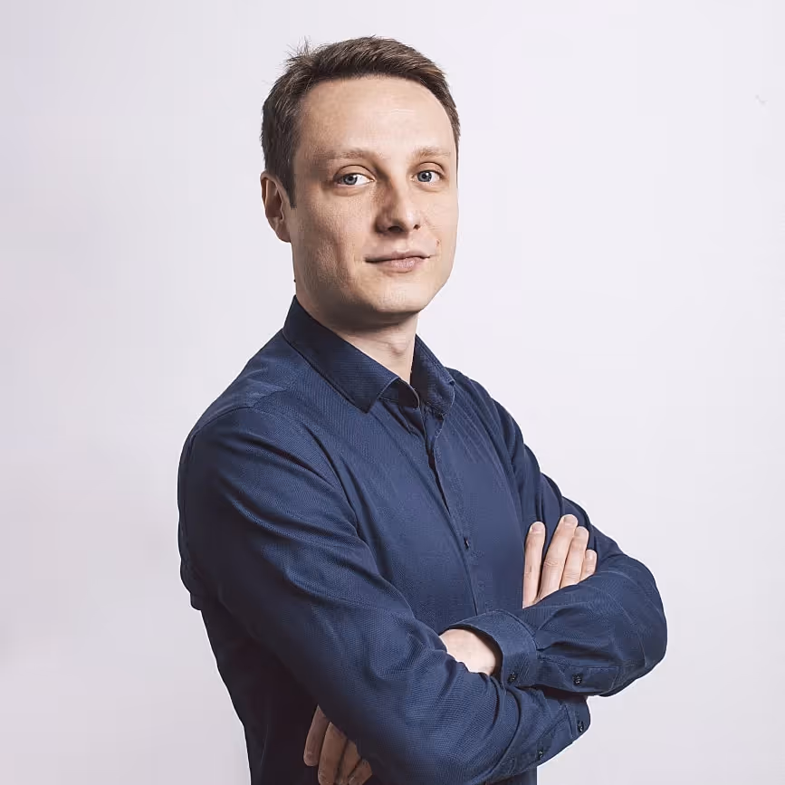 Man with short brown hair wearing a dark blue buttoned shirt with arms crossed, looking confidently at the camera.