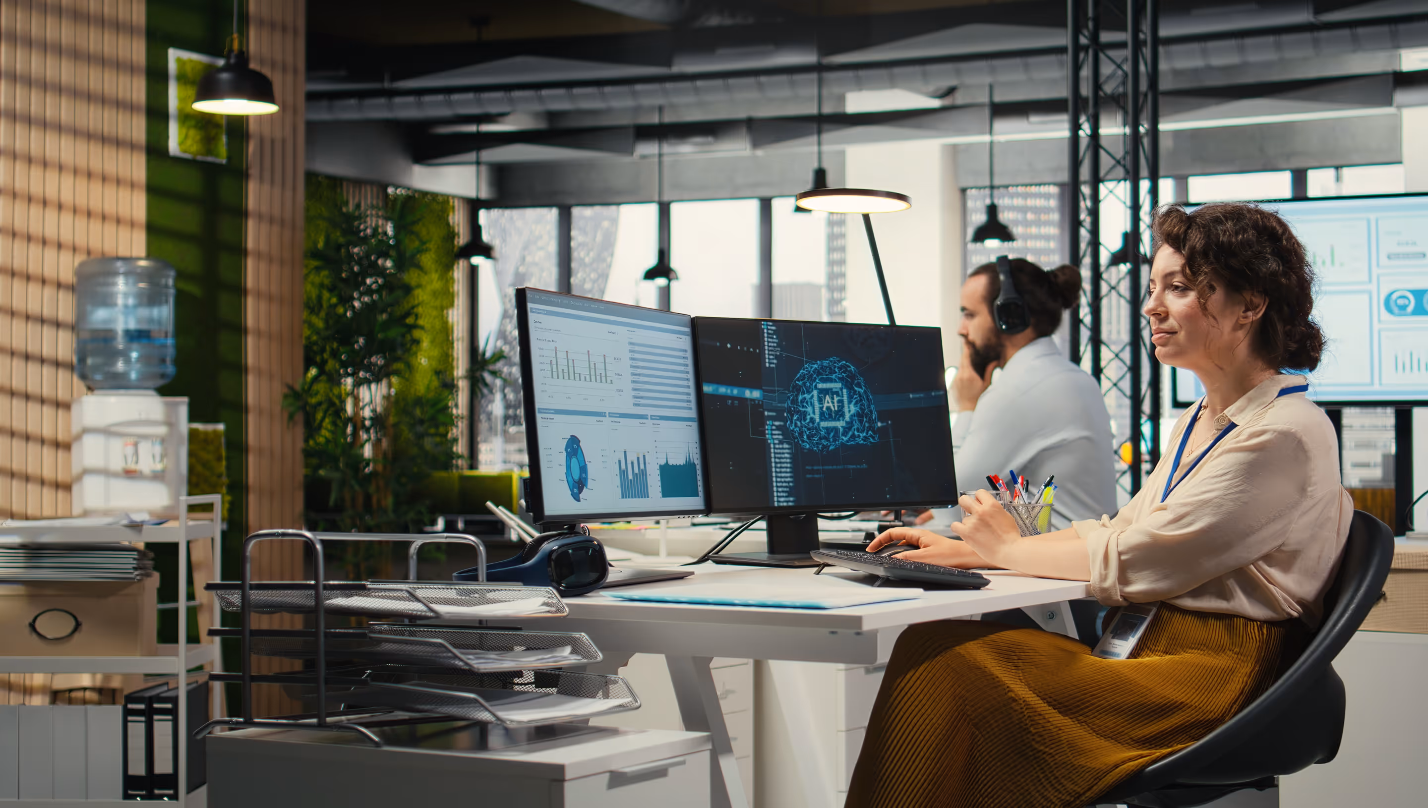 Two people working at a shared desk with dual monitors displaying AI brain graphics and data charts in a modern office.