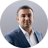 Portrait of a man with short dark hair wearing a gray blazer and white shirt against a light gray background.