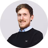 Smiling man with short brown hair and beard wearing a black sweater and blue collared shirt against a light background.