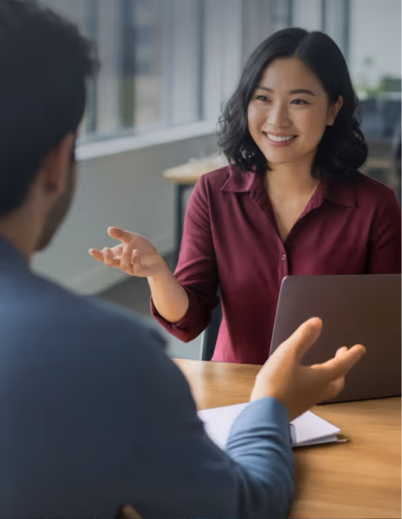 Smiling woman in a burgundy shirt gestures while talking to a man across a desk with a laptop.