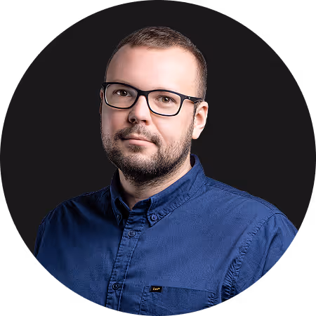 Man with glasses and trimmed beard wearing dark blue buttoned shirt on black background.