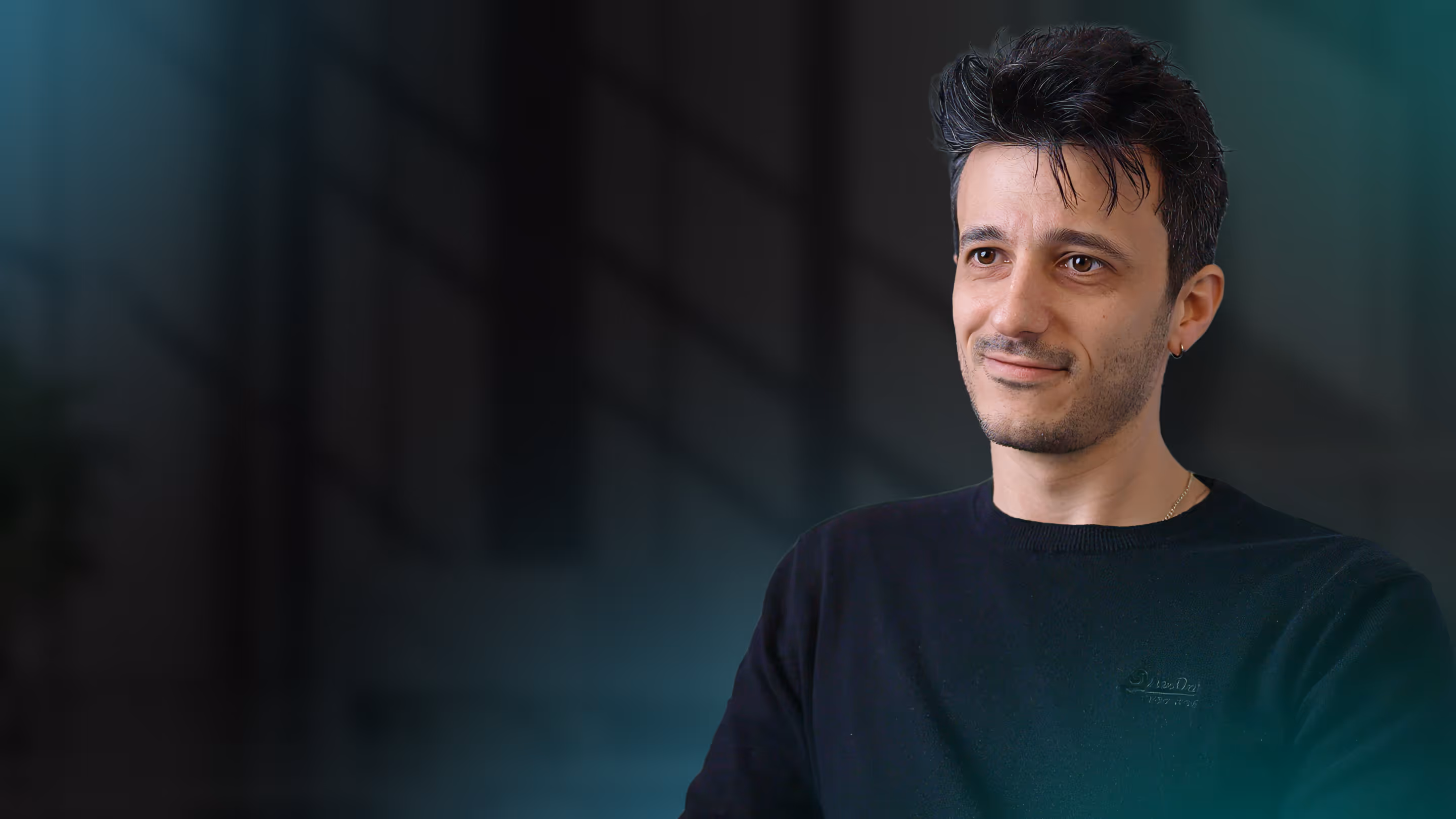 Man with short dark hair and earring wearing a black sweater, smiling slightly, against a dark background.