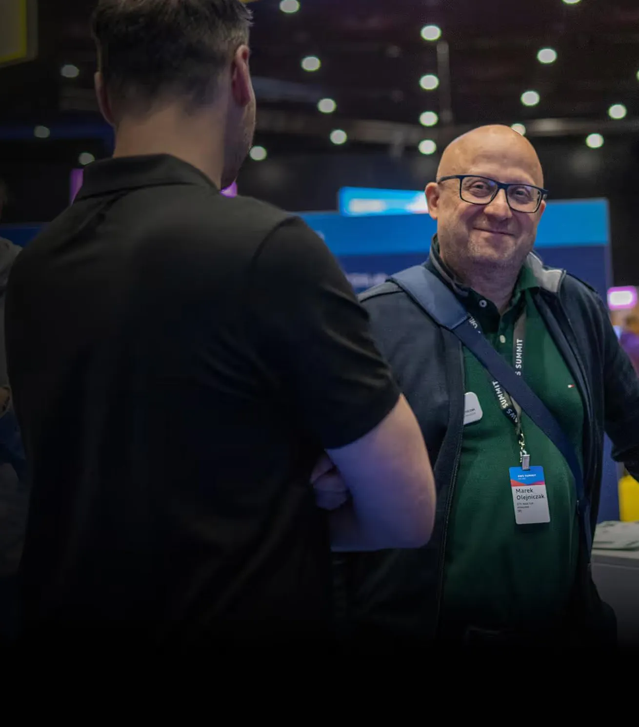 Two men interacting at a conference or event, one facing away wearing a black shirt, the other bald with glasses and a blue jacket, smiling with a name badge that reads Marek Olejniczak.