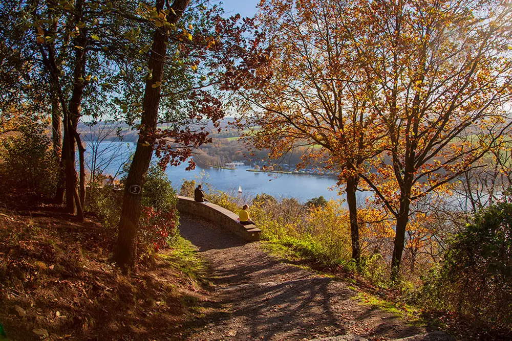 Zwei Personen sitzen auf einer Steinmauer, Blick über den See, herbstliche Farben im Hintergrund, ruhige Wasseroberfläche