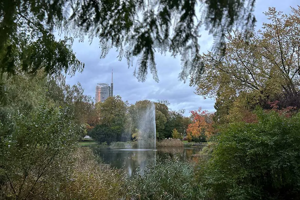 Ententeich mit sprudelnder Wasserfontäne umgeben von altem Baumbestand vor blauem Himmel, Stadtgarten Essen