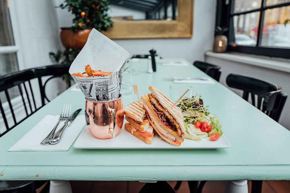 Süsskartoffelpommes in Spitztüte im Kupferbecher und Chicken Sandwiches mit Salat auf calypsofarbenen Holztisch.