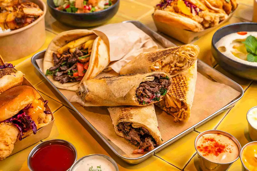 Zwei Tabletts mit Beef- und Chicken-Shawarma, Burgern und Pommes, ergänzt durch verschiedene Saucen und Dips auf einem gelb gefliesten Hintergrund