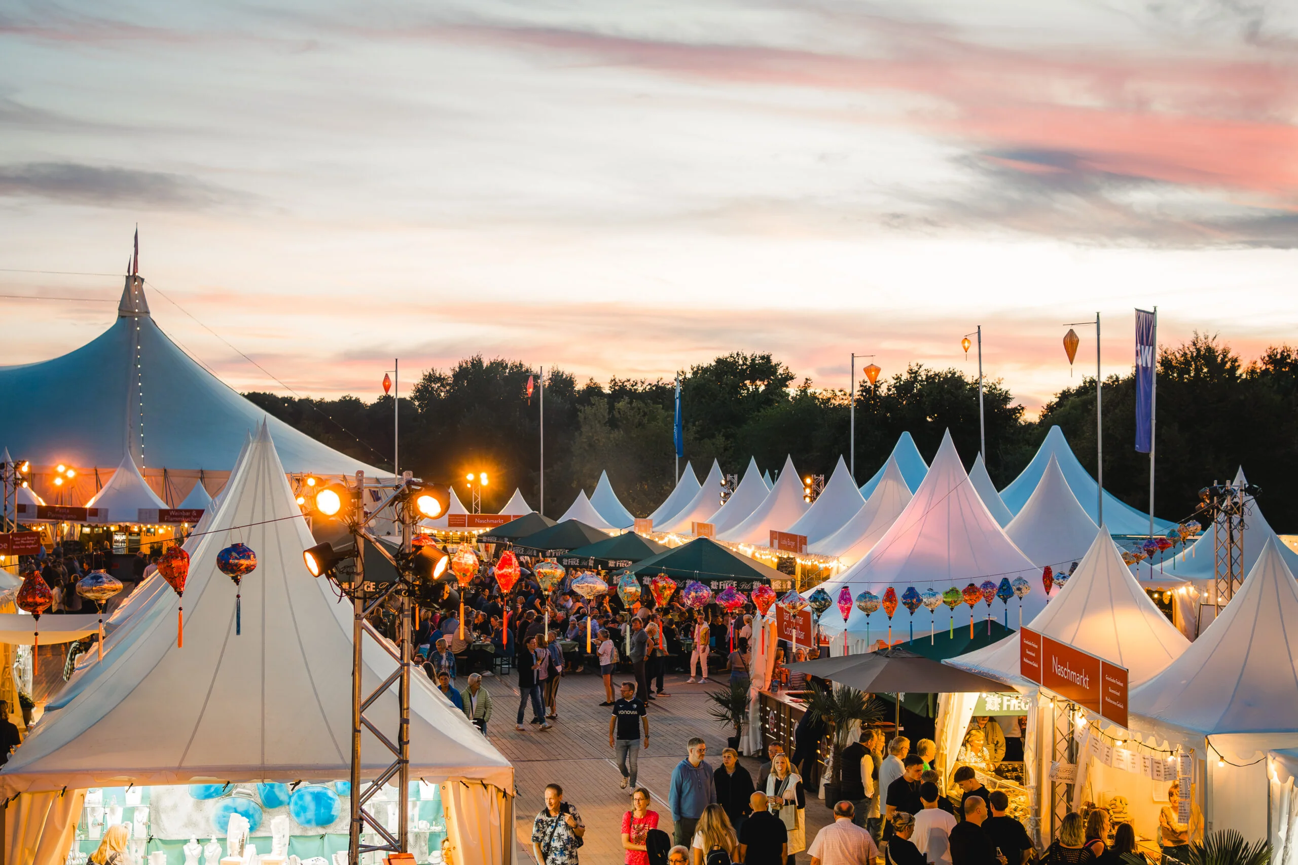Open-Air-Festival mit einer weißen Zeltstadt, Marktständen, bunten Lampions und vielen Menschen bei Sonnenuntergang