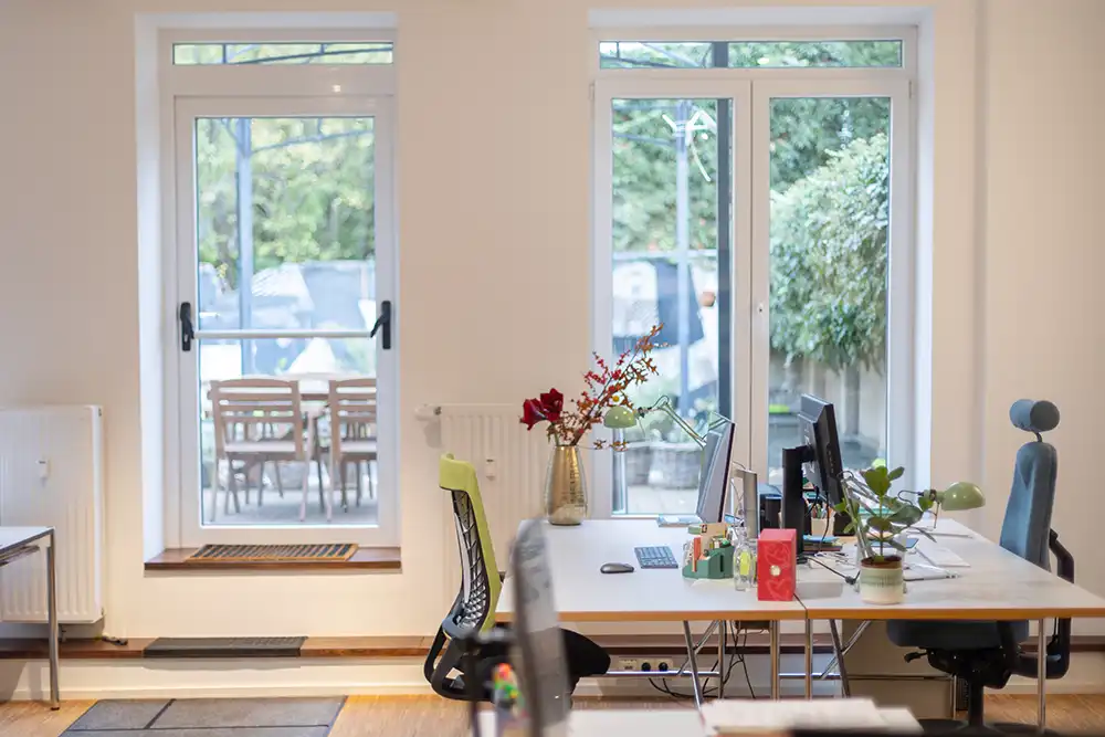 Heller Büro-Arbeitsplatz im Coworking Space mit modernen Schreibtischen und Blick durch bodentiefe Fenster in einen grünen Garten