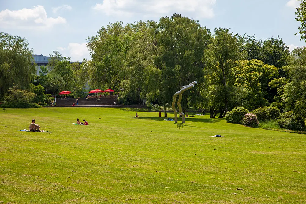 Mehrere Personen und Paare sitzen und liegen entspannt beim Picknick auf der großen Liegewiese im Essener Grugapark neben einer modernen Skulptur.