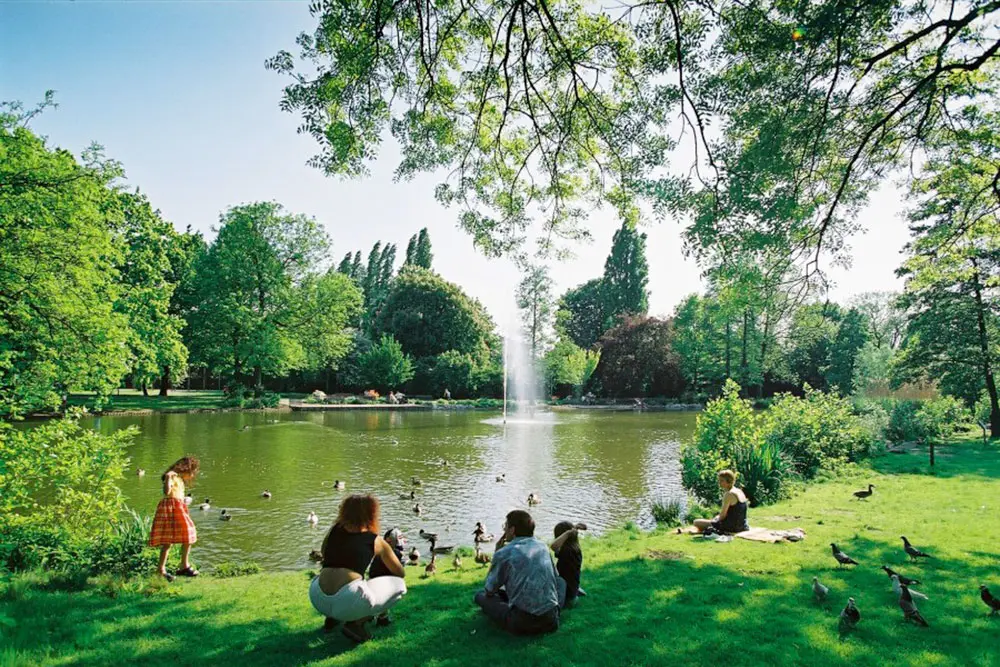 Besucher:innen sitzen im Gras am Ufer eines großen Teichs mit Wasserfontäne im historischen Kaiser-Wilhelm-Park in Essen-Altenessen.