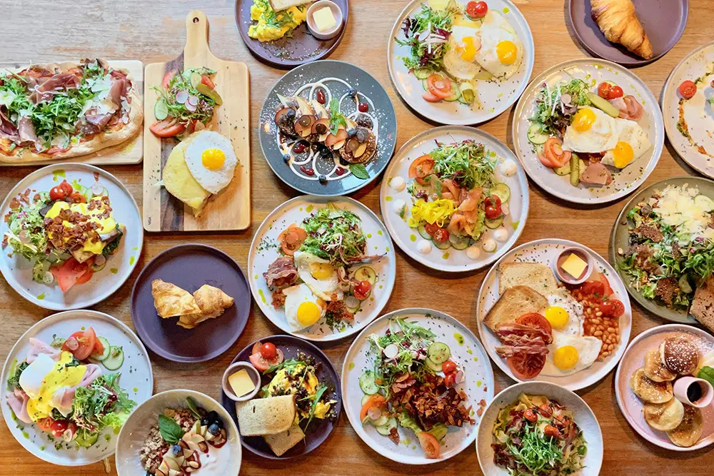 Ein Holztisch von oben fotografiert, reich gedeckt mit verschiedenen Frühstückstellern wie Spiegeleiern, Pancakes, Croissants und Salaten in einem Café in Essen.