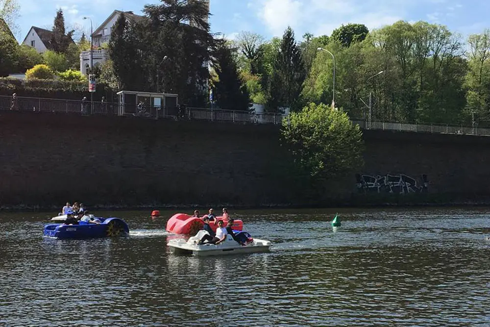 Besucher:innen in bunten Tretbooten auf der Ruhr in Essen-Kettwig vor einer bewaldeten Uferpromenade im Sommer.