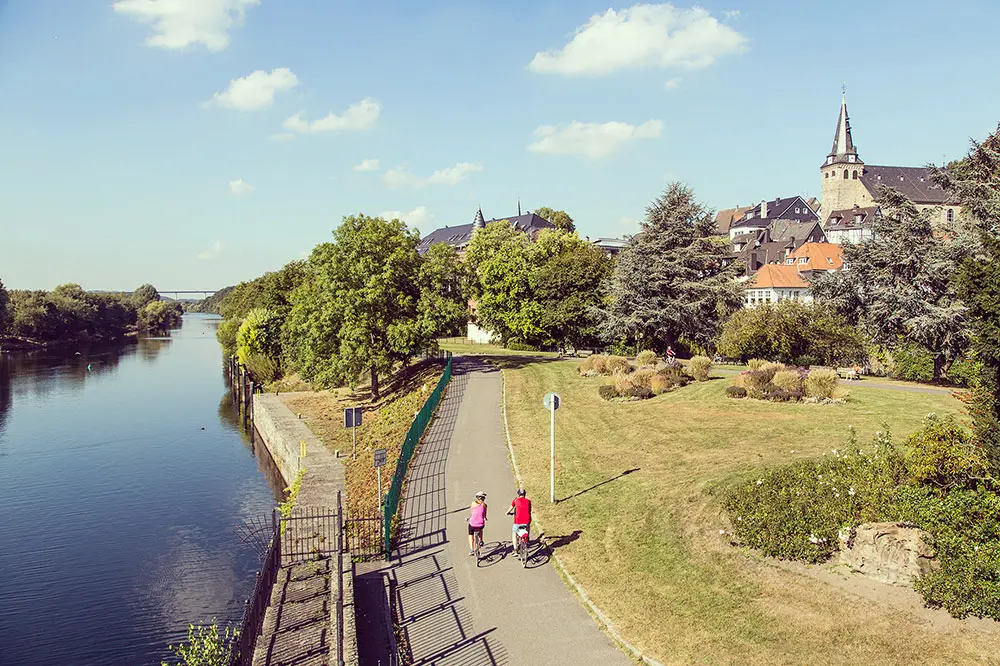 Zwei Radfahrer:innen auf dem RuhrtalRadweg in Essen-Kettwig direkt an der Ruhr vor der historischen Altstadt.