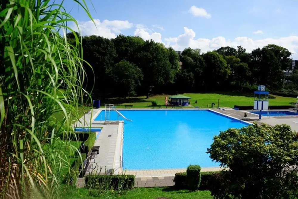 Sonntag-Veranstaltungs-Tipp in Essen, Blick auf das leere Schwimmbecken im Freibad Kettwig bei Sonnenschein, mit einer kleinen Rutsche, blauem Sprungturm und dichter Bewaldung im Hintergrund.
