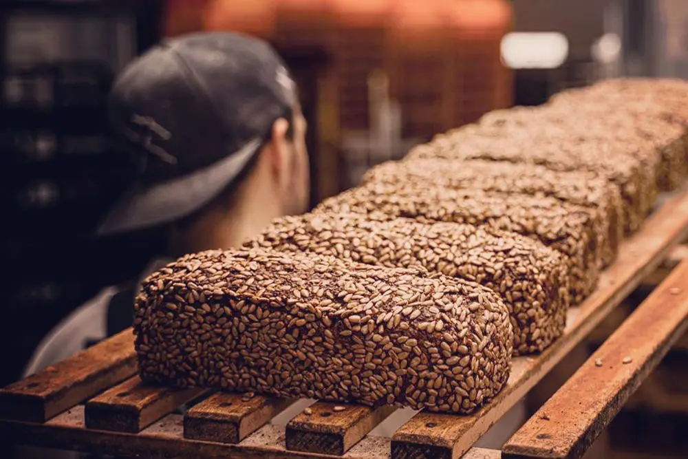 Ein Bäcker in der Backstube der Bäckerei Rabauken in Essen hinter einer Reihe frisch gebackener Vollkornbrote mit Sonnenblumenkernen.