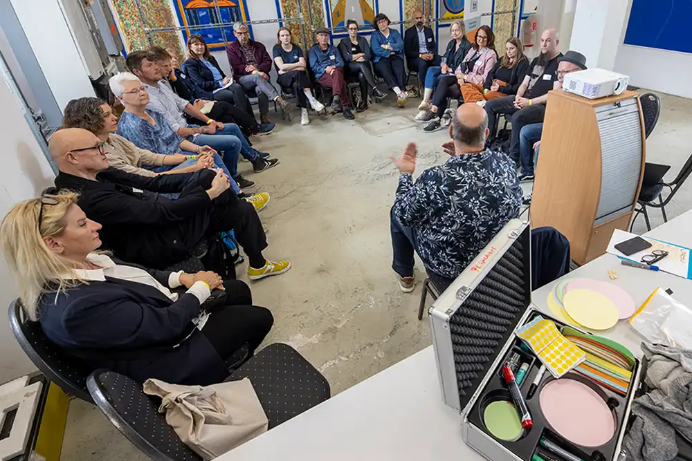 Eine Gruppe von Leuten sitzt im Kreis auf Stühlen in einem hellen Raum mit großen Fenstern und nimmt an einem Workshop teil.