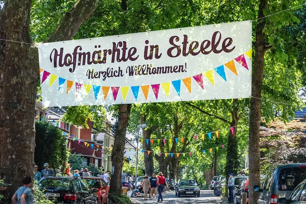Ein großes Willkommensbanner mit Wimpelkette überspannt eine grüne Wohnstraße bei den Hofmärkten in Essen-Steele.