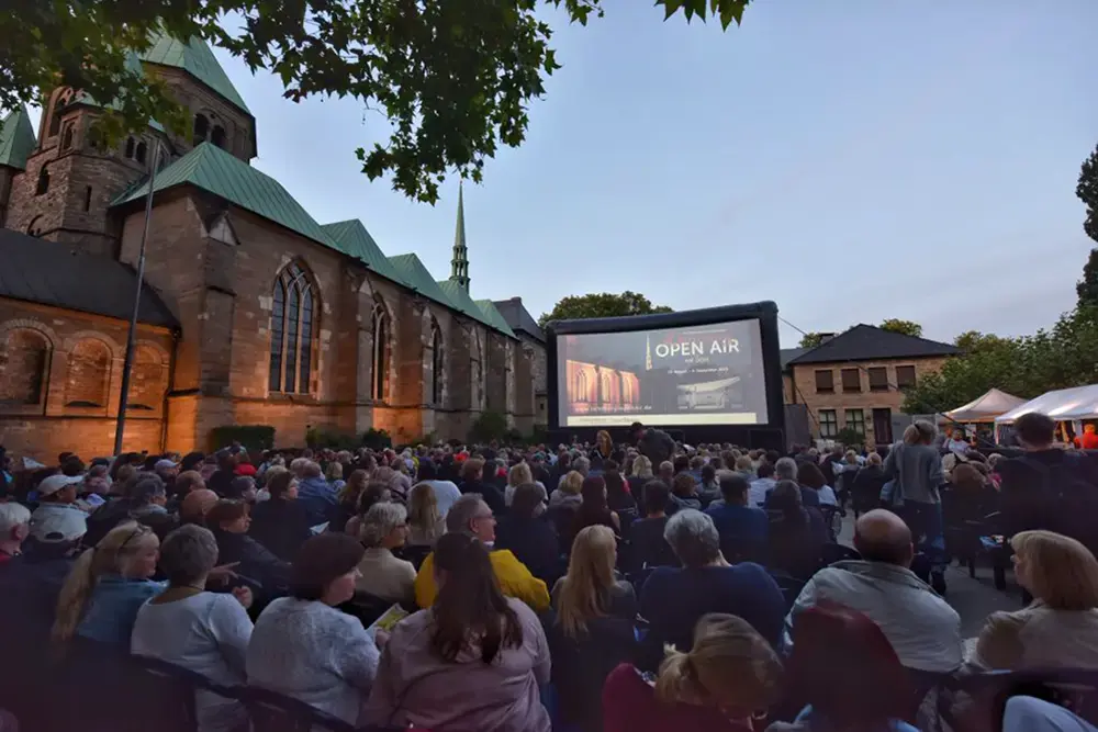 Zahlreiche Zuschauer:innen sitzen vor einer großen Open-Air-Kinoleinwand neben dem beleuchteten Essener Dom beim Lichtburg Open Air Kino in den Sommerferien.