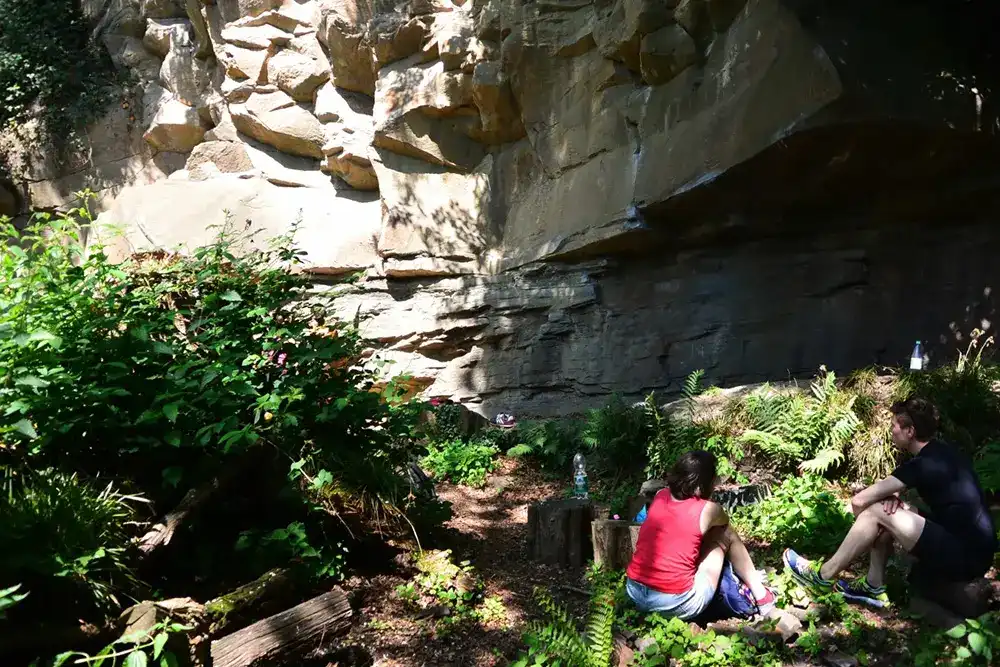 Zwei Personen sitzen im Schatten vor der imposanten Naturkletterwand Nangijala in Essen während eines Ausflugs in den Sommerferien.
