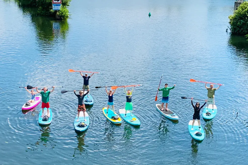 Eine Gruppe von Menschen steht gut gelaunt mit erhobenen Paddeln auf Stand-up-Paddle-Boards (SUP) auf der Ruhr in Essen-Kettwig bei einer Tour von Glück auf SUP.