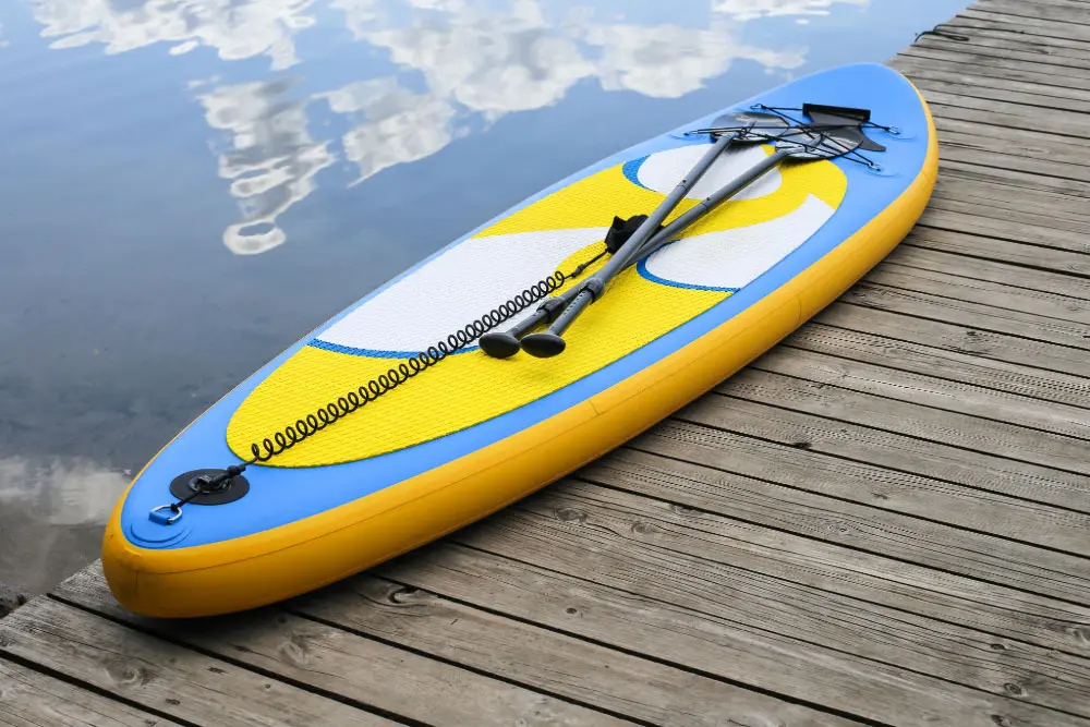 Ein gelb-blaues SUP-Board mit Paddel liegt auf einem Holzsteg am Wasser beim Seaside Beach Essen am Baldeneysee.