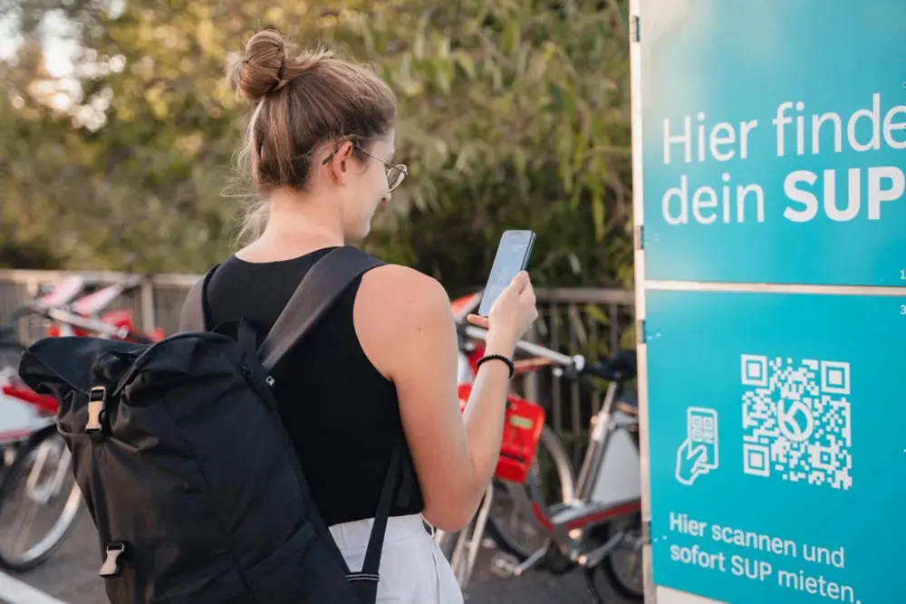 Eine junge Frau scannt mit ihrem Smartphone einen QR-Code an einer blauen Verleihstation, um ein SUP-Board am Campingpark KNAUS in Essen am Baldeneysee zu mieten.