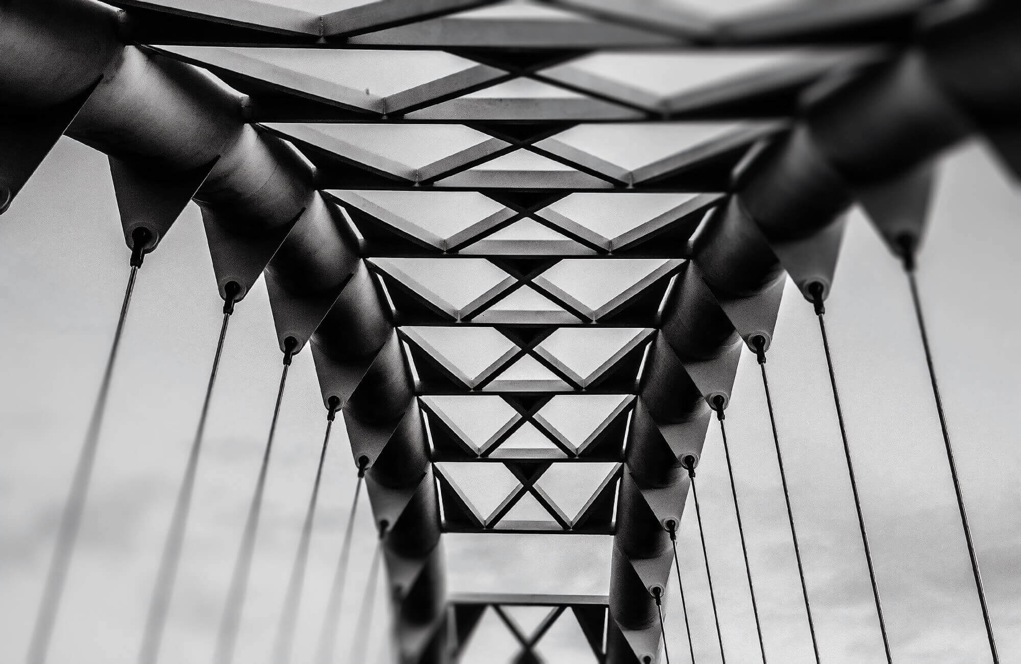 Photo looking up through the geometric steel framework and suspension cables of a bridge against a sky.