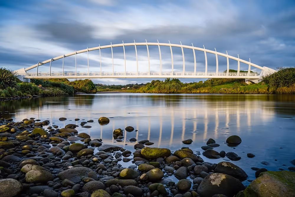 Modern white bridge over a calm river with rocks and greenery.